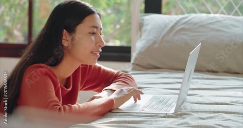 Asian Teenage Girl in Red Shirt Using Laptop on Bed, 
 in Cozy Bedroom with Warm Morning Sunlight, Side View.