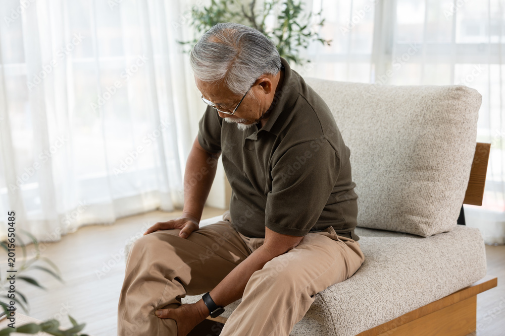 Fototapeta premium Senior man sitting on comfortable sofa in bright living room feeling distressed and experiencing sharp pain in knee joint suffering from painful arthritis ache while rubbing leg at home