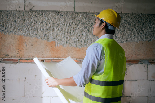 Asian male civil engineer in safety vest and hard hat inspecting blueprints at construction site.