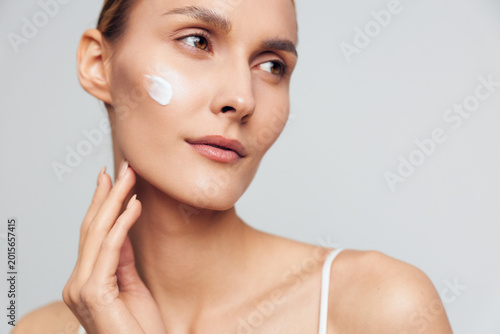 Cuadro en lienzo Young woman with a dewy complexion applies cream to her cheek in a studio portrait, with soft lighting highlighting smooth skin and a calm vibe