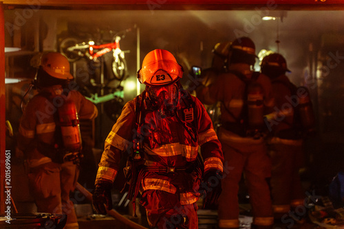 Slika na platnu Firefighter in full turnout gear exiting burning building with smoke and other firefighters in the background