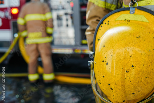 Fotografia Yellow firefighter helmet covered in soot and melted tar with a firefighter looking over a fire engine pump panel in the background
