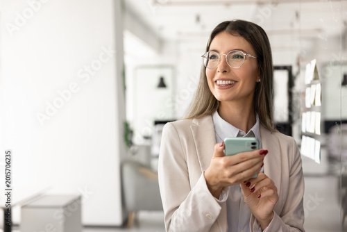 Photography Confident young businesswoman stands at modern workplace, holding smart phone, smiles with satisfaction, checking important work emails, exchanging messages