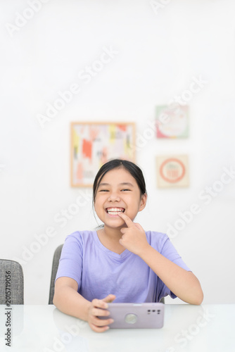 Asian child smile white teeth in dental clinic or young kid girl and people happy holding playing smartphone or mobile phone and point to tooth for show present permanent teeth and baby tooth vertical