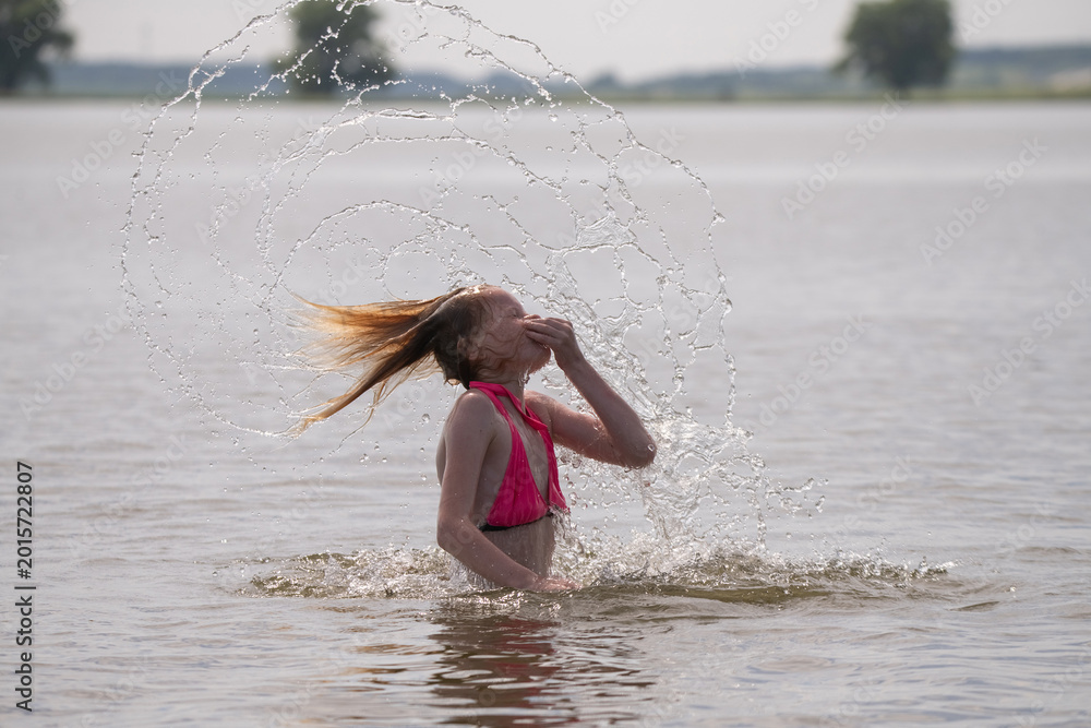 Naklejka premium Female swimmer in pink bikini splashes water while flipping hair back in a lake, with trees and a clear sky visible in the background