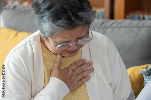 Bilde på lerret Senior asian woman sitting on indoor couch holding chest experiencing pain distr