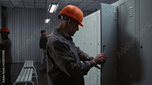 Male Industrial Worker Unlocks Metal Cabinet, Checks Wristwatch, and Removes Hard Hat, Changing Clothes While Friendly Interacting With Coworkers in Modern Locker Room. Professional Factory Routine.