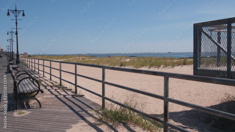 Fototapeta premium Coney Island beach promenade in Brooklyn, New York, United States. Wooden boardwalk near retro luna park. Benches on ocean coast, sandy beach. Waterfront summer holiday in NYC, USA. Outdoor recreation