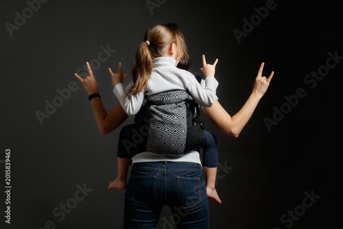 Mother and little girl in back carrier showing rock gestures with hands, studio shot from behind.