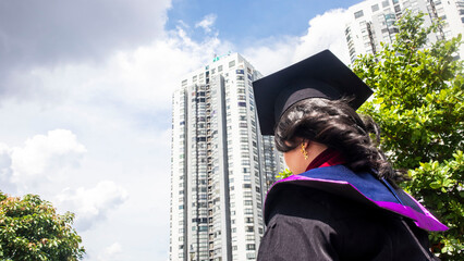 A girl who is happy with her graduation, she is wearing a graduation cap and graduation gown with a background of tall buildings.