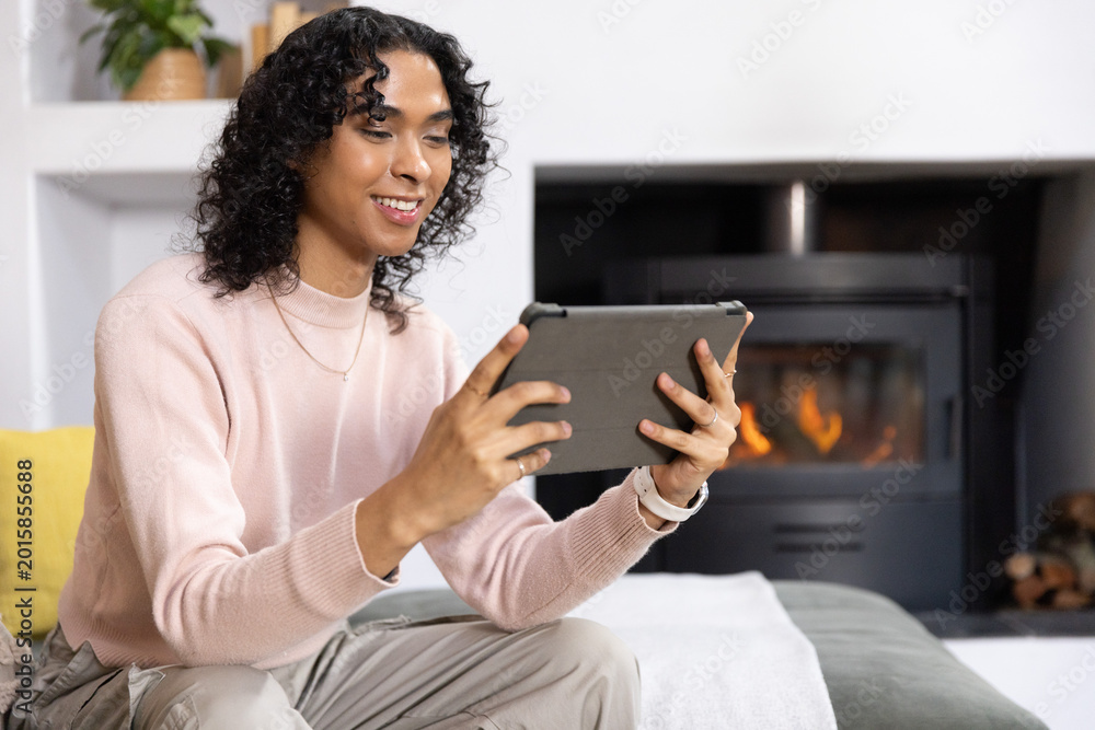 Obraz premium Non-binary adult sitting on sofa in living room holding tablet, in pink sweater, fireplace burning
