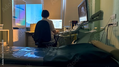 Female medical technician in ultrasound exam room, seated at imaging workstation with monitors and control panel, calm clinical mood, healthcare diagnostics setting