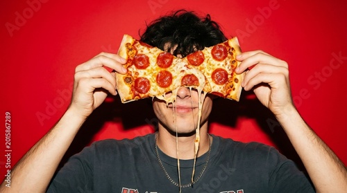 Young man holds a piece of pepperoni pizza over his eyes while posing against a solid red studio background to emphasize his love for junk food in this stylized and modern lifestyle photography shot.