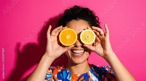 Woman holding slices of orange and lemon over her eyes like glasses while laughing in front of a saturated hot pink wall, creating a fun and colorful visual display of fresh citrus energy.