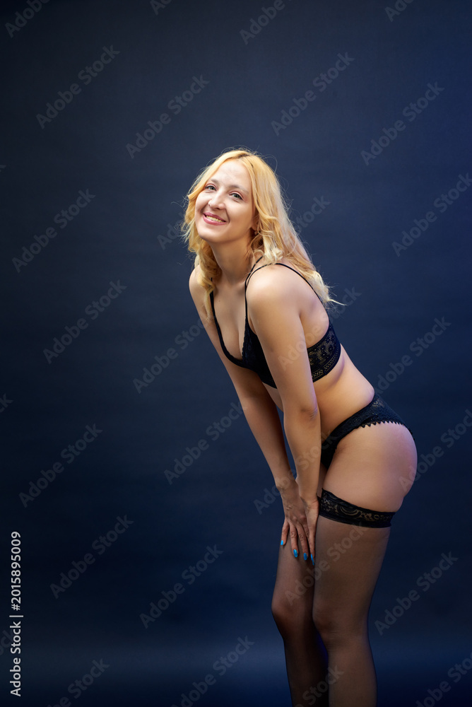Fototapeta premium A young, sexy woman in black lingerie poses against a dark blue background. She smiles gently. Her pose exudes charm and attractiveness. This is a concept for a fashion and beauty magazine.