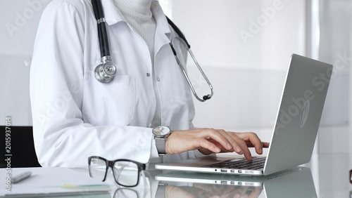 Foto Woman doctor wearing white coat and stethoscope, typing on laptop in clinic office, using modern medical technology