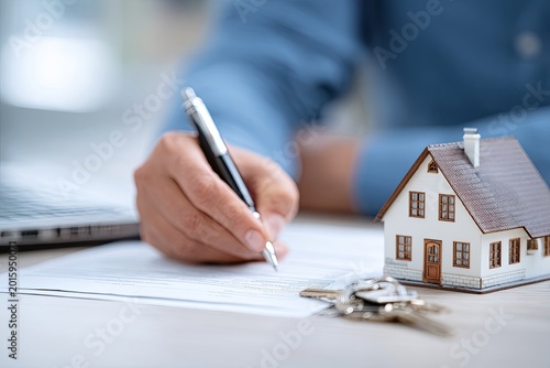 Person signing documents with house model and keys nearby