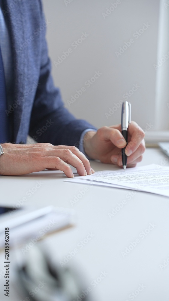 Fototapeta premium Closeup of businessman signing a contract with elegant pen, wearing a blue suit and a wristwatch, sitting at a white desk in office. Business people concept