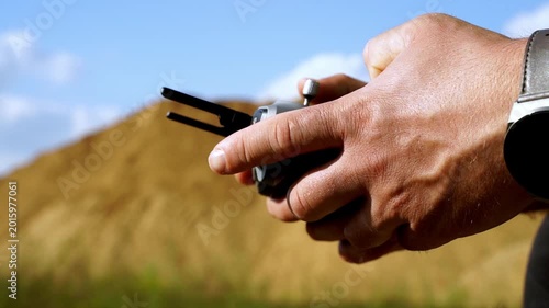 Hands grip remote, focused on drone control. Blue sky, golden earth, soft focus background. Steady hands suggest calm, precise operation. Outdoor drone piloting, likely for survey or photography