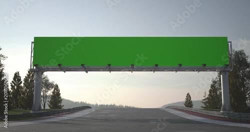 Race cars drive under a green billboard on a racing track, wide view with asphalt, horizon and forest landscape in daylight