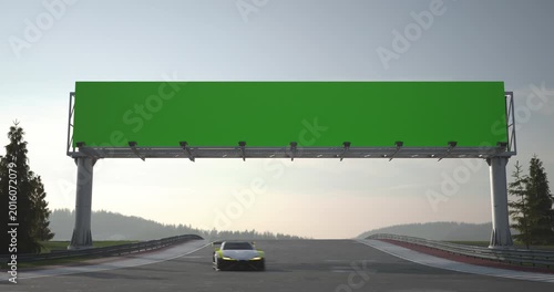 Race cars drive under a green billboard on a racing track, wide view with asphalt, horizon and forest landscape in daylight