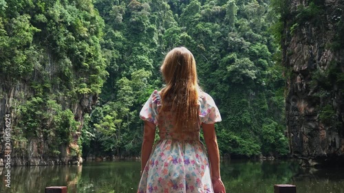 Young woman in floral dress looking at tropical lagoon surrounded by lush limestone cliffs and dense jungle during daytime. Back view of traveler exploring scenic nature in Southeast Asia.