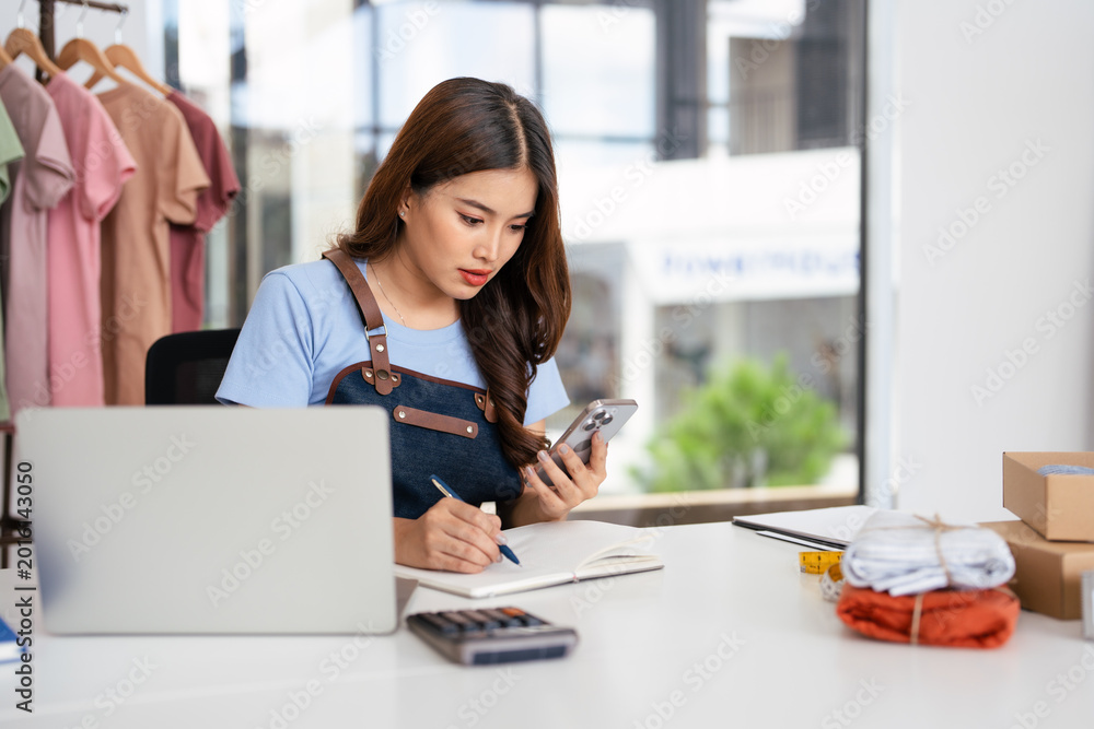 Fototapeta premium Small business owner wearing an apron, jotting down notes on a notepad while working on cell phone.