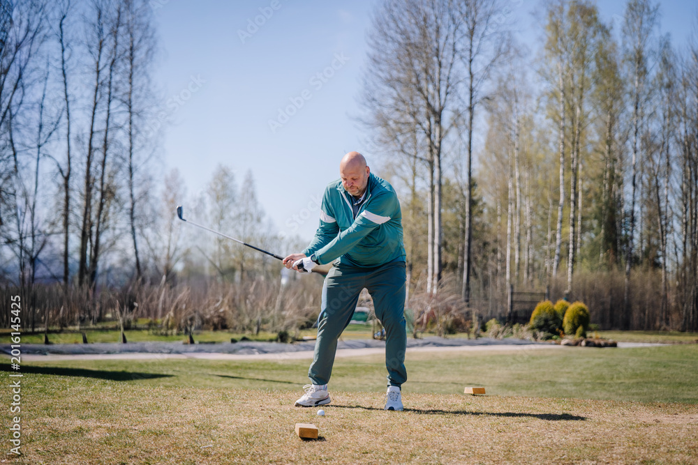 Obraz premium Senior man in teal athletic wear swings golf club on sunny day at outdoor golf course with trees and grass in the background