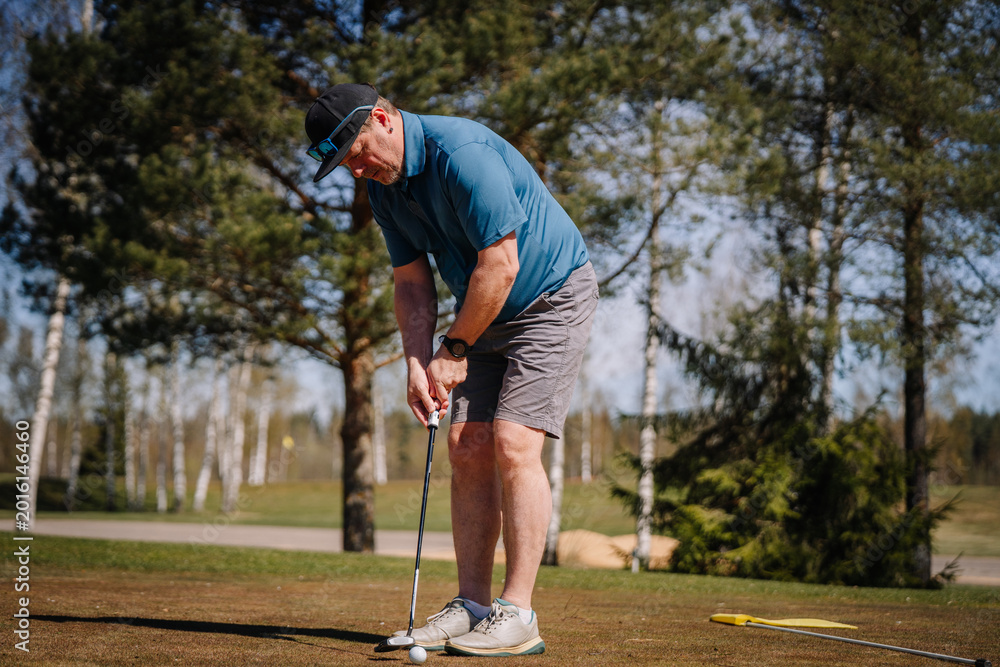 Obraz premium Senior man putting golf ball on green during sunny day, surrounded by trees and open space on a golf course