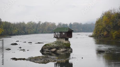 Lonely cottage on rock in middle of Drina river near Bajina Basta in Serbia, autumn colors on forest on riverbanks and fog obscures mountains in background, popular tourist attraction