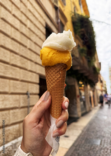 Person holding a colorful yellow gelato in a cone on a street in Italy, with historic buildings in the background reflecting Italian culture and cuisine. Yellow gelato in hand.
