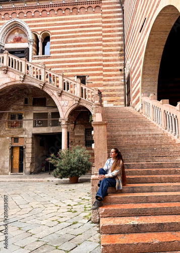 Young woman sitting on marble steps of medieval Palazzo della Ragione in Verona. Striped brick facade, Gothic arches and a fresco create a stunning backdrop.