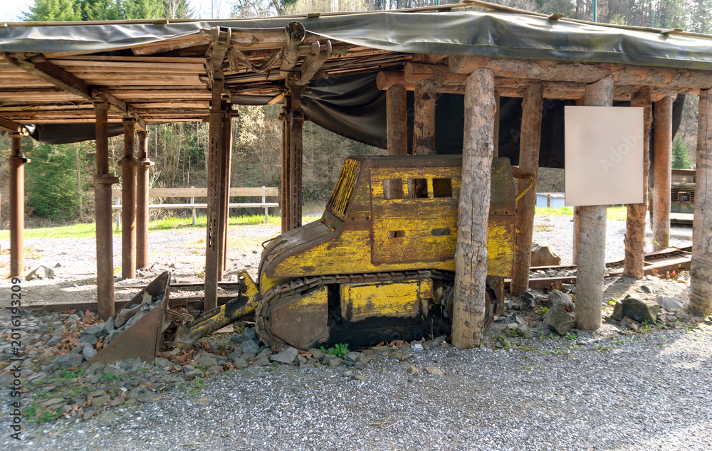 Fototapeta premium Weathered yellow bulldozer with front blade and crawler tracks stands under a rough wooden shelter with rocks around it. The scene shows abandoned construction machinery, quarry work, industrial decay