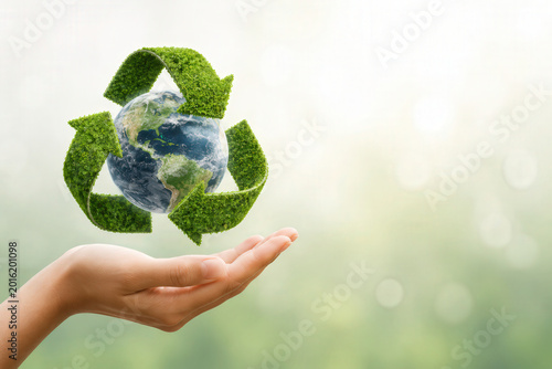 A hand holds the earth with a green recycling symbol above it. The background is blurred and suggests an outdoors setting with light coming through. This shows the idea of caring for the planet.