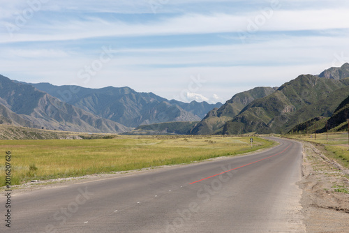 Empty asphalt road of the Chuysky Trakt running through a wide mountain valley with green meadows and dramatic ridges, Altai Republic, Russia, sunny day