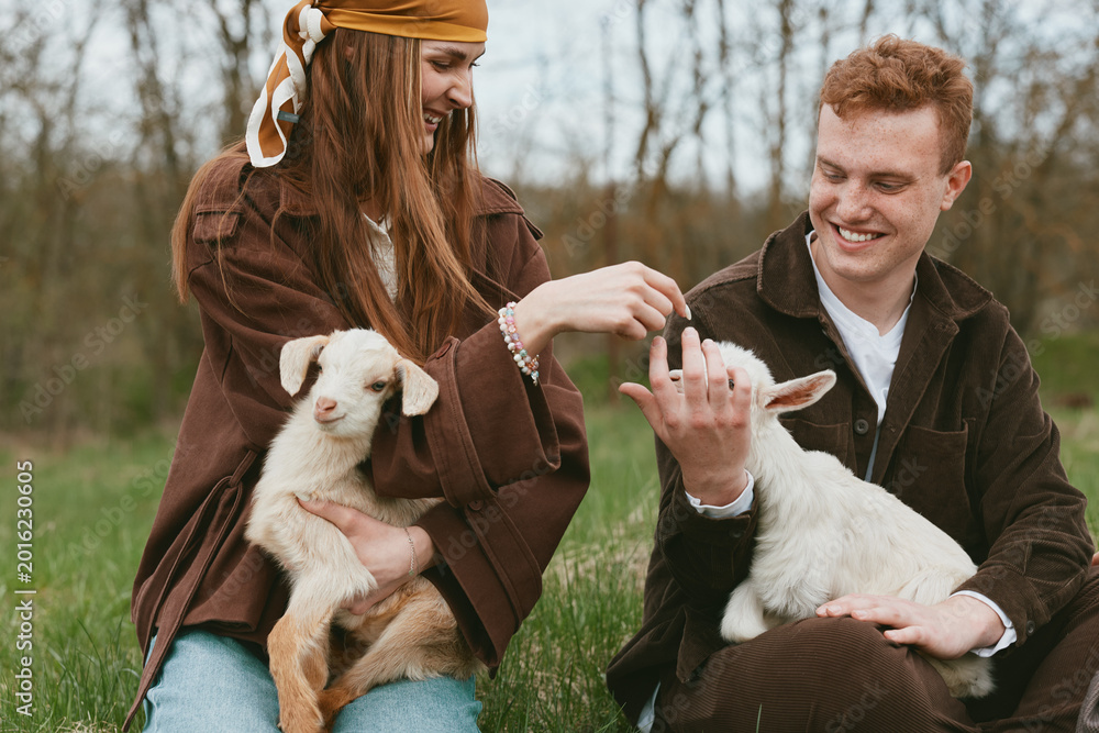 Naklejka premium Cheerful couple outdoors with a goat kid, gentle interaction in a sunlit meadow, rustic vibe, warm colors, natural lighting, candid portrait of bonding and playful affection with animals
