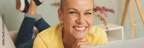 Картината върху платно Smiling woman dressed in yellow T-shirt and jeans lying on sofa with laptop looking at camera