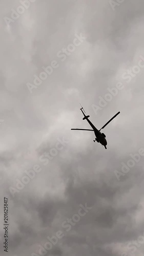 Military helicopter silhouette flying through overcast gray cloudy sky above national flag.