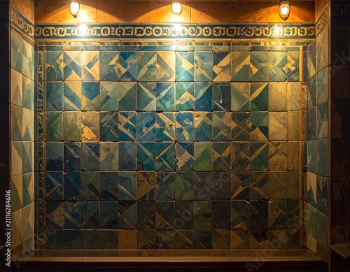 Illuminated vintage wall adorned with an intricate mosaic of aged blue and green geometric ceramic tiles, highlighting traditional craftsmanship and rich texture