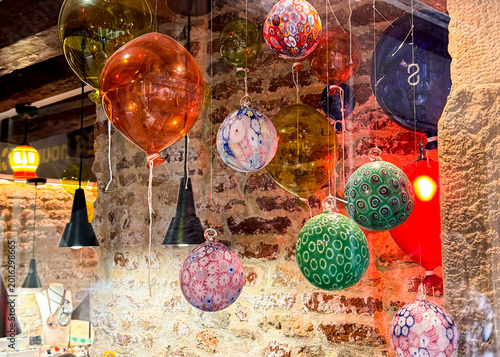 Colorful handmade Murano glass ornament balls hanging against a rustic brick wall in a Venice shop. Warm atmospheric lighting highlights the millefiori patterns and vivid colors.