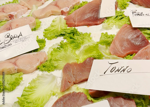 Fresh tuna and swordfish fillets displayed on lettuce leaves with handwritten Italian labels at a traditional Venice fish market. Authentic local seafood trading scene.