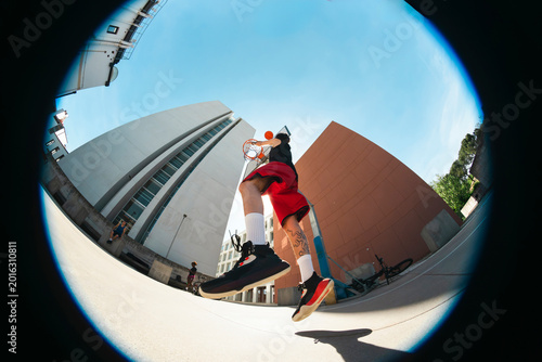 A young man dunks a basketball on an outdoor court. Other players and spectators are in the background. A wide-angle lens captures modern architecture under a bright sky.