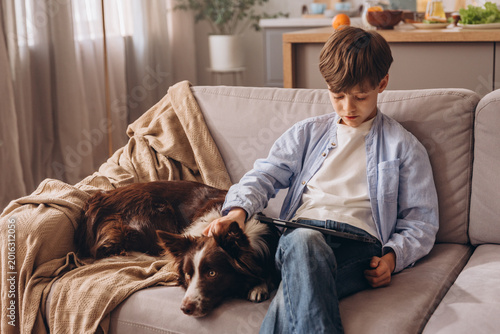 Boy using digital tablet and petting his dog while relaxing on a sofa
