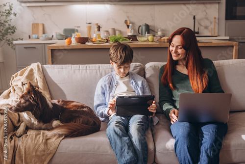 Mother working on laptop and watching her son use a digital tablet at home
