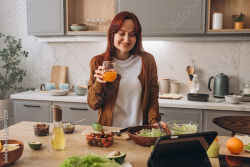 Cheerful woman drink taste orange juice while watching cooking lesson via digital tablet in kitchen
