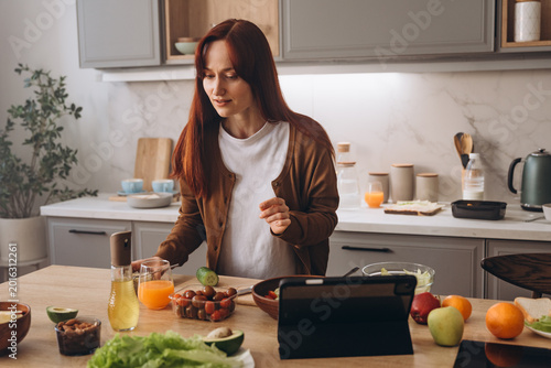 Young redhead woman cooks salad as watches online master class at home
