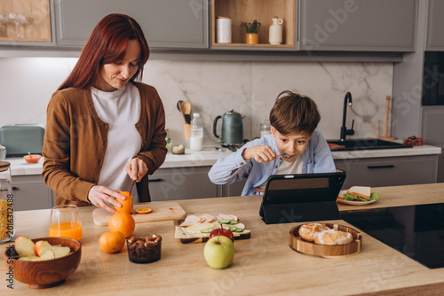 Konstfotografi Mother cooks breakfast as preteen son eats breakfast and watches video via digit