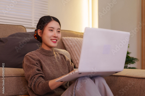 фотографія young Asian woman using laptop computer working from home sitting on sofa smilin