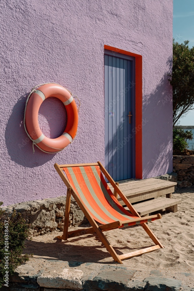 Fototapeta premium Deck chair on sandy shore beside purple building doorway