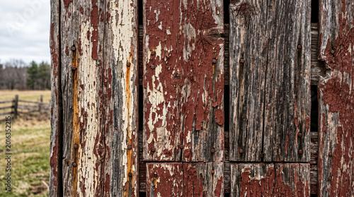 Weathered barn wood with peeling red paint reveals intricate textures of age and decay, a rustic facade against a blurred rural landscape